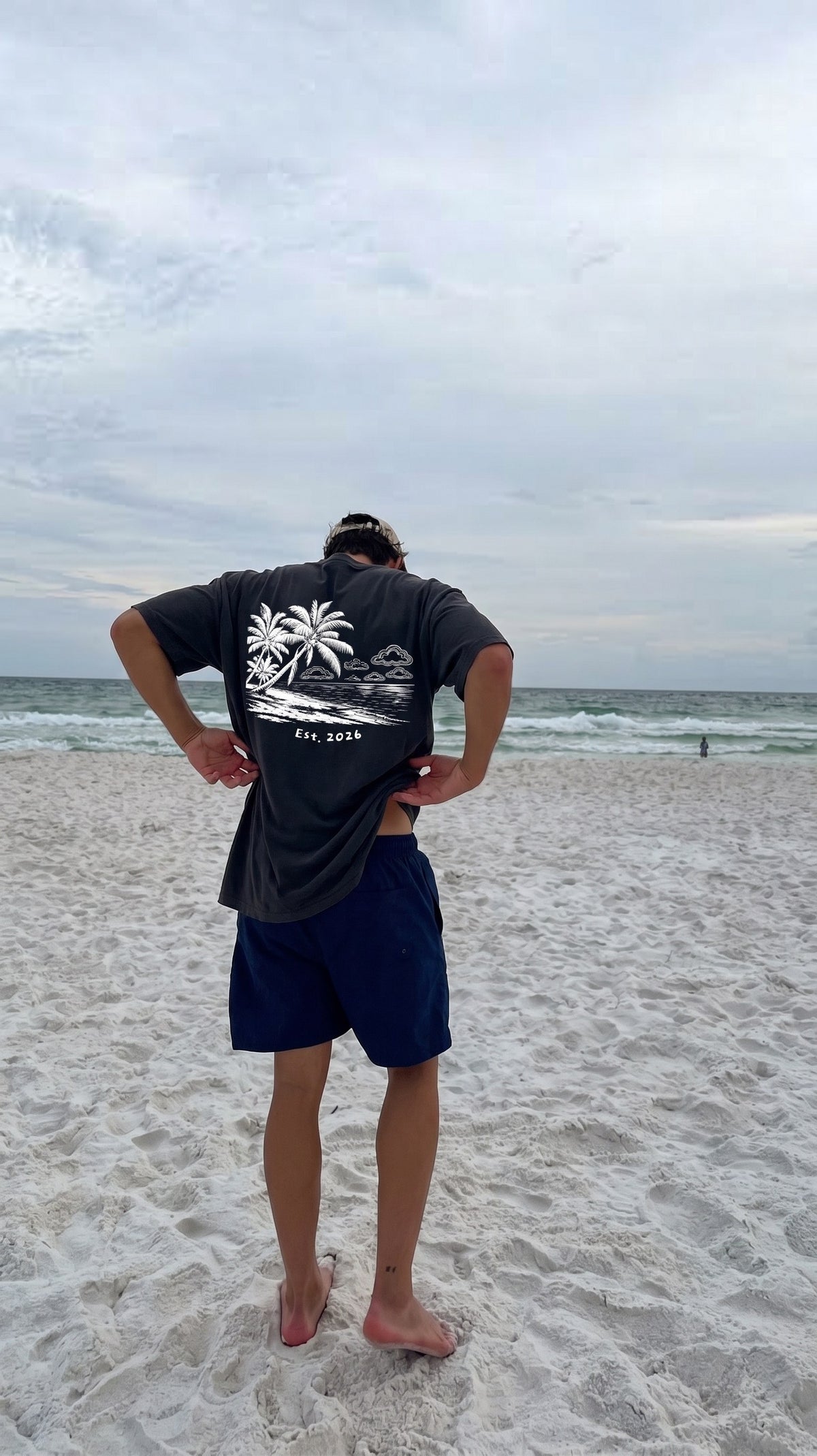 Man on a white-sand beach in the Palm Beach tee, palm-and-clouds back print